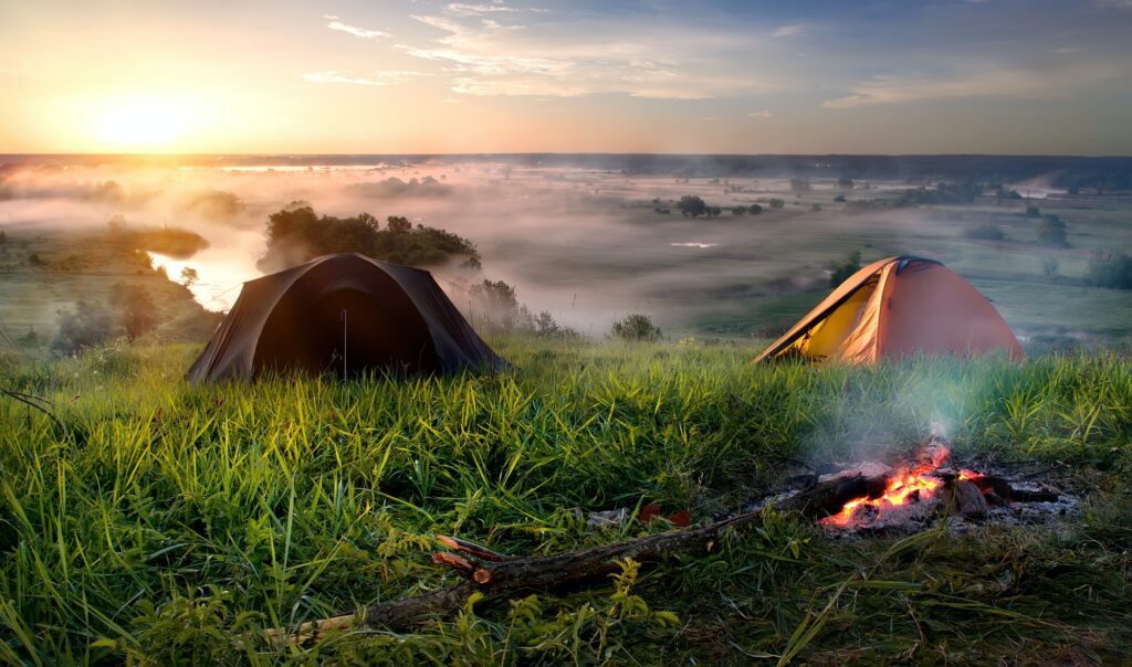 MMA Resorts Mekkarai Two tents sit on a grassy hillside overlooking a misty valley at sunrise, with a small campfire burning in the foreground and a river winding through the landscape in the distance.