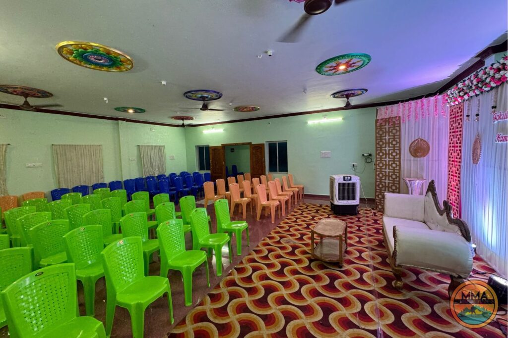 MMA Resorts Mekkarai A decorated event hall with rows of green, blue, and peach plastic chairs facing a stage area with a white sofa, colorful ceiling decorations, patterned carpet, and draped backdrop.