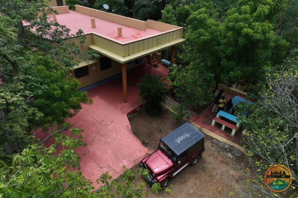 MMA Resorts Mekkarai Aerial view of a house with a red roof and patio surrounded by trees. A red jeep is parked on the driveway, and several people are gathered near a blue table outside. An MMA logo appears in the bottom right corner.