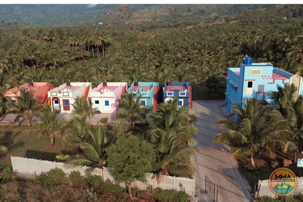 Aerial view of colorful houses and a tall blue building surrounded by palm trees and greenery, with mountains visible in the background. Reminiscent of Western Ghats resorts Tenkasi, the property is enclosed by a white fence.